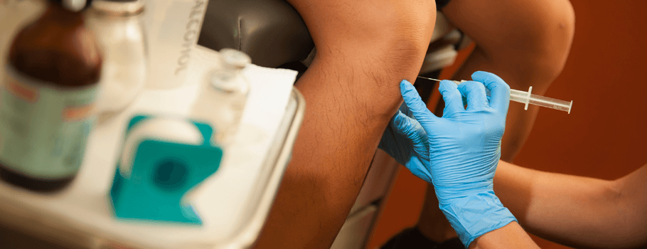 A healthcare professional wearing blue gloves is administering a vaccine or injection into a patient's arm, with the focus on the syringe and the point of injection.