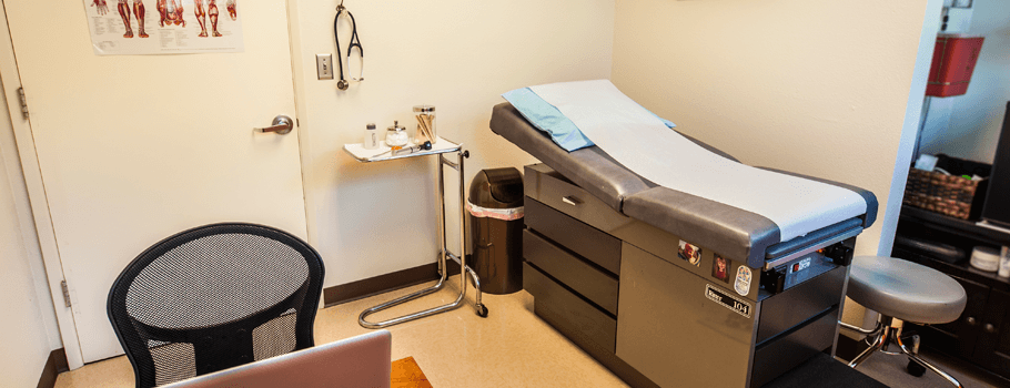 An examination room with a medical examination table, a stool, a laptop on a desk, and anatomical posters on the wall, ready for the next patient.