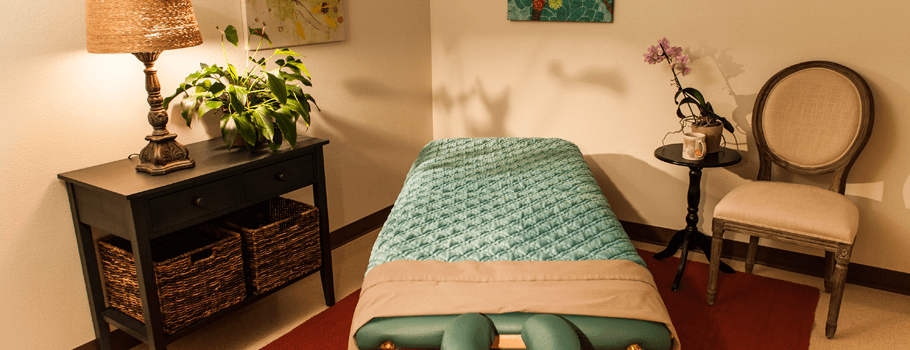 A cozy and warmly lit therapy or massage room with a comfortable table, soft lighting from a table lamp, a potted plant and elegant furniture promoting a relaxing atmosphere.