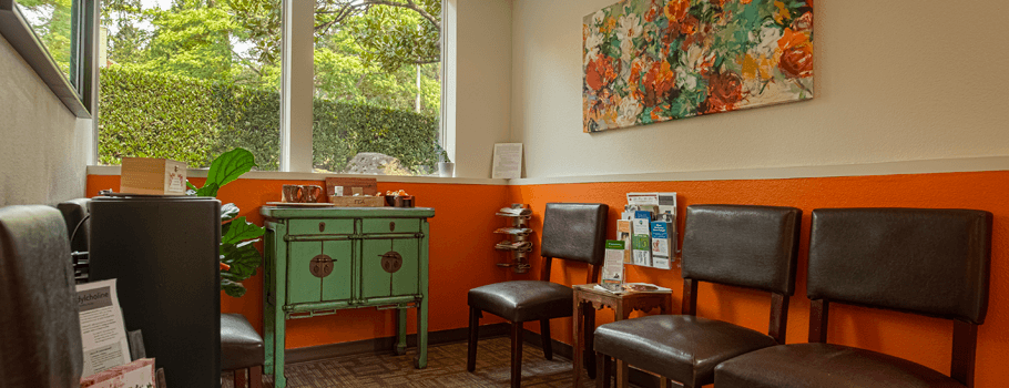 A cozy waiting area with warm orange walls, comfortable seating, a decorative floral painting, and a wooden cabinet with informational brochures, creating a welcoming atmosphere.