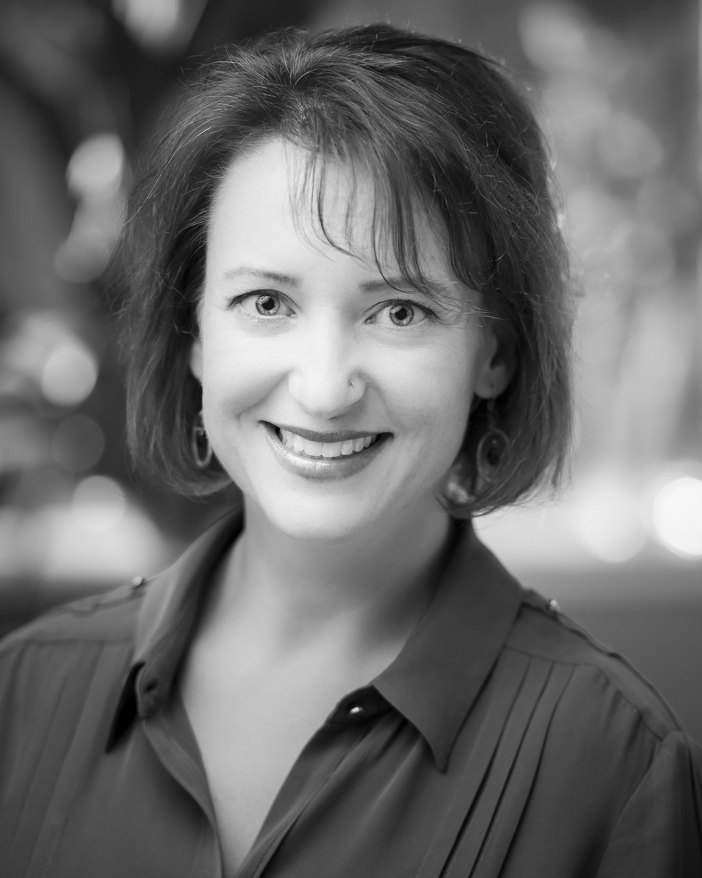 A black and white portrait of a smiling woman with short hair, standing in front of a softly blurred background that suggests lights or bokeh. her friendly expression and direct gaze create a warm and approachable atmosphere.