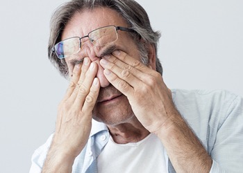 Elderly man rubbing tired eyes while wearing glasses, possibly experiencing eye strain or fatigue.