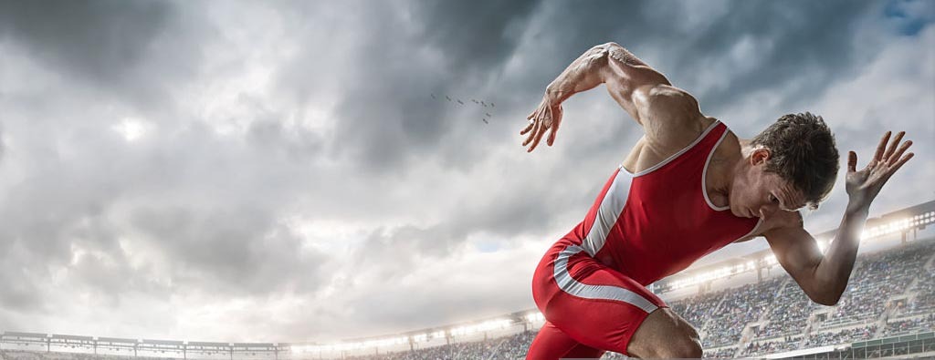 A mid action image of a professional male athlete sprinting from starting blocks on an outdoor athletics running track. The runner is in a generic outdoor floodlit stadium full of spectators under a dramatic stormy sky. The sprinter wears a red body suit and running spikes during a practice session.