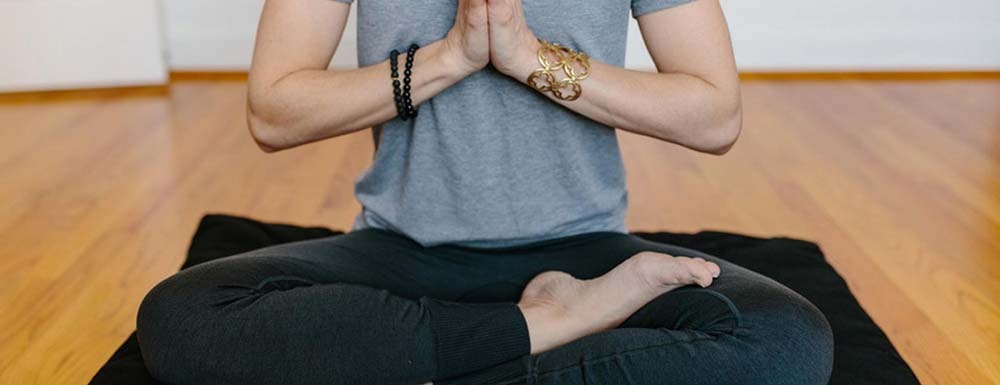 A person in a gray t-shirt and black pants sits cross-legged on a yoga mat, performing a meditation pose with their hands pressed together in an anjali mudra.