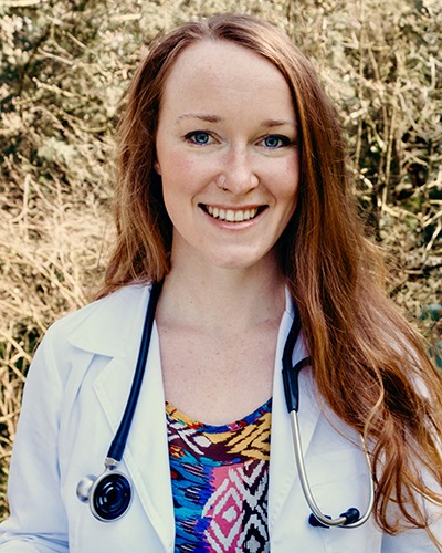 A healthcare professional poses with a friendly smile, wearing a lab coat with a stethoscope around the neck, standing before a backdrop of natural greenery.