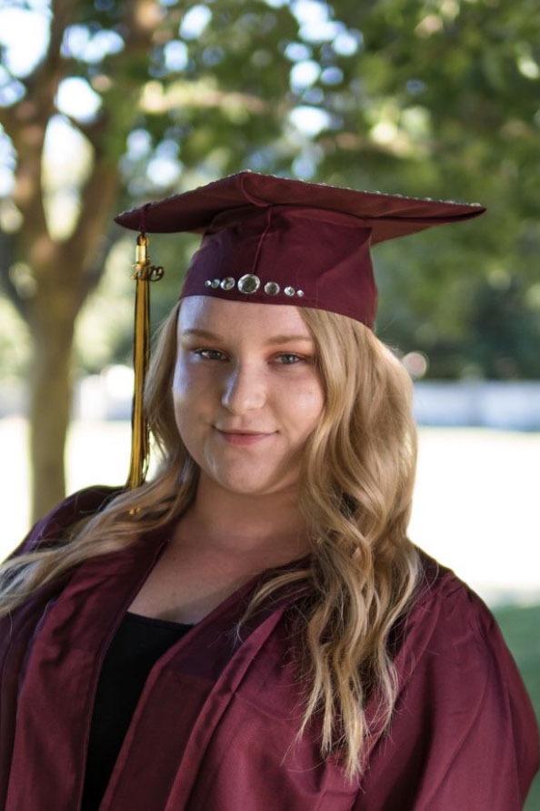 A proud graduate wearing a maroon cap and gown, smiling with a backdrop of greenery, celebrating a significant academic milestone.