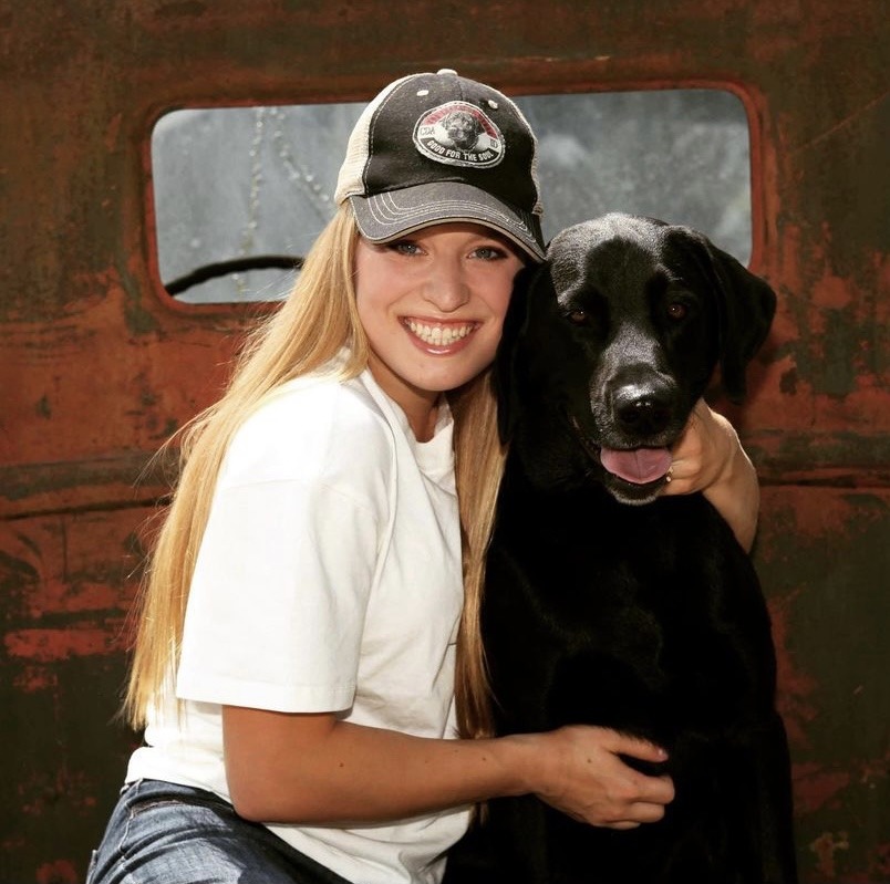 A smiling young woman wearing a baseball cap hugging a happy black dog, with both sitting in front of a rusty vehicle.