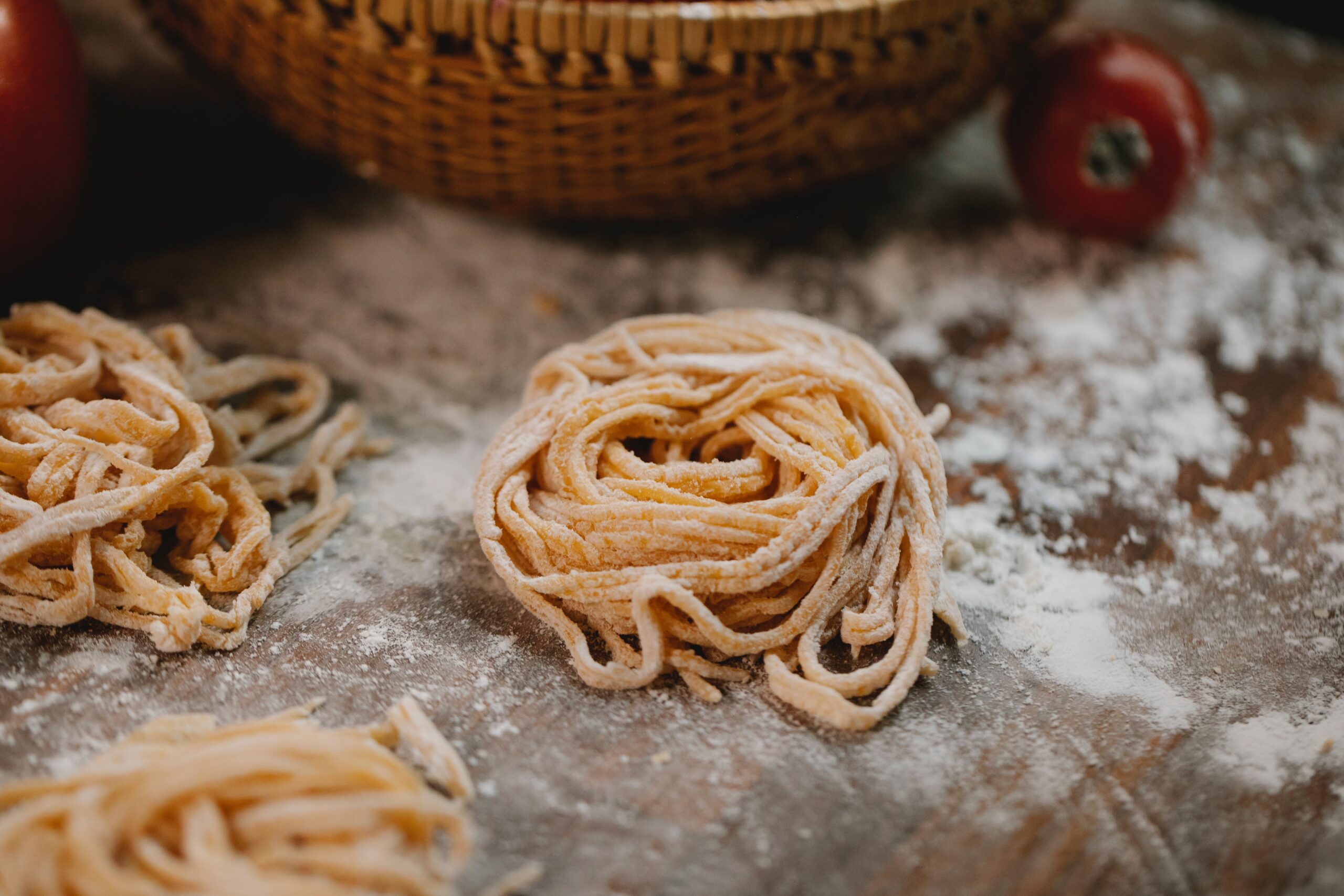 Fresh homemade pasta nest on a floured wooden surface with tomatoes and a basket in the background.