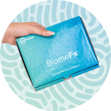 Hand holding a BiomeFx gut microbiome test kit against a blue-green background.