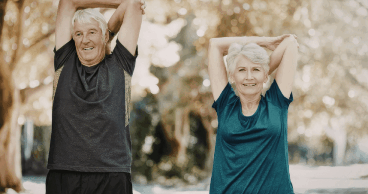 Senior couple standing outdoors, arms stretched overhead and back, smiling while doing a stretch in a park.