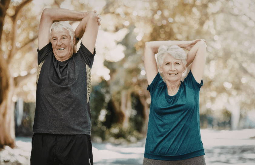 Senior couple standing outdoors, arms stretched overhead and back, smiling while doing a stretch in a park.