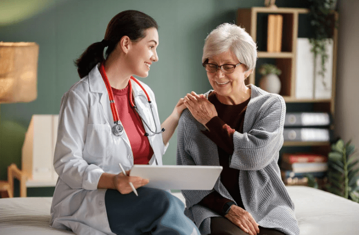Doctor in a white coat with a stethoscope sits with an elderly woman, smiling during a medical consultation in a cozy room with a clipboard/tablet nearby.