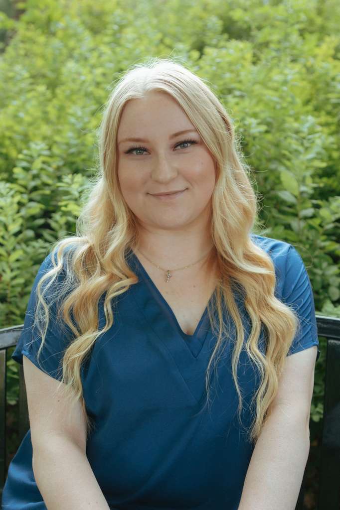 Portrait of Britney, medical assistant at Lake Oswego Health Center, smiling with long blonde hair wearing blue scrubs outdoors, green foliage in the background.