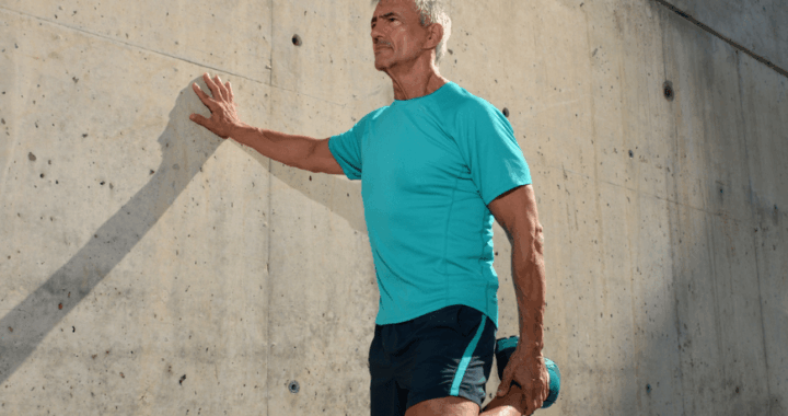 Senior man performing a standing quadriceps stretch against a concrete wall outdoors before exercise, wearing athletic clothing and warming up for fitness, mobility, and flexibility training in sunny weather.