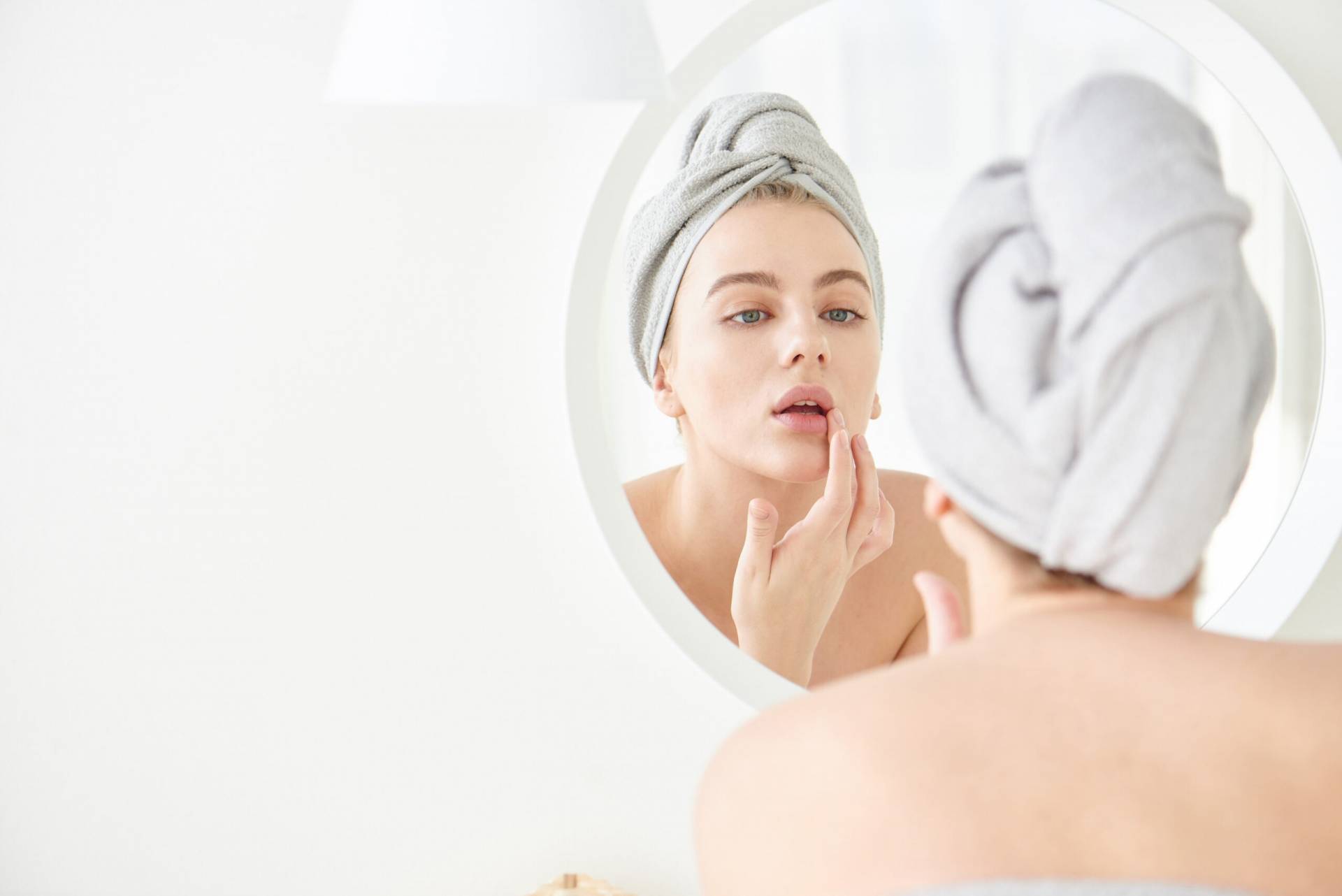 Portrait of  young women with  towel on head in white bathroom looks and touches her face in the mirror and enjoys youth and hydration. Natural beauty, home care for problem skin